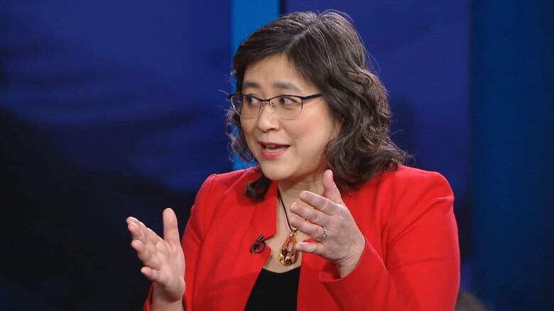 A woman wearing a red blazer and yellow pendant necklace gestures while speaking. 