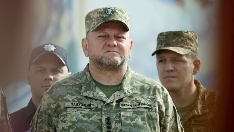 A stocky man with a chin beard is shown in military fatigues with a serious look on his face.