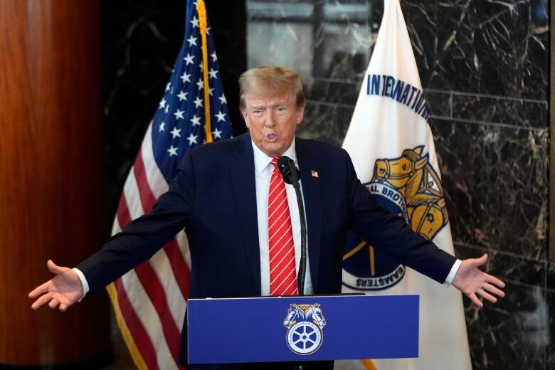 A man in a suit and tie speaks at a podium and extends his arms out, in front of a pair of flags.