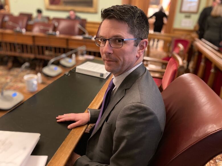 A man with short, dark hair and glasses, wearing a grey suit, white button-down shirt and purple tie, sitting at a long desk, looking at the camera over his left shoulder.