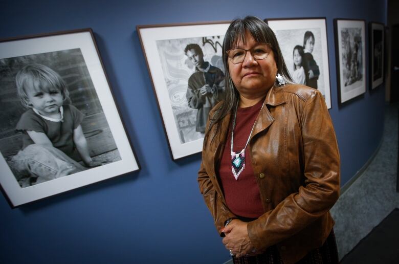 A woman in long black hair stands near a blue wall. Black and white photos of children are in frames and hanging on the wall