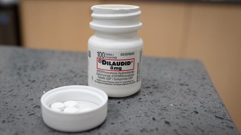 A bottle labeled dilaudid, a pharmaceutically made version of hydromorphone, is open with white pills in the lid, on a counter.