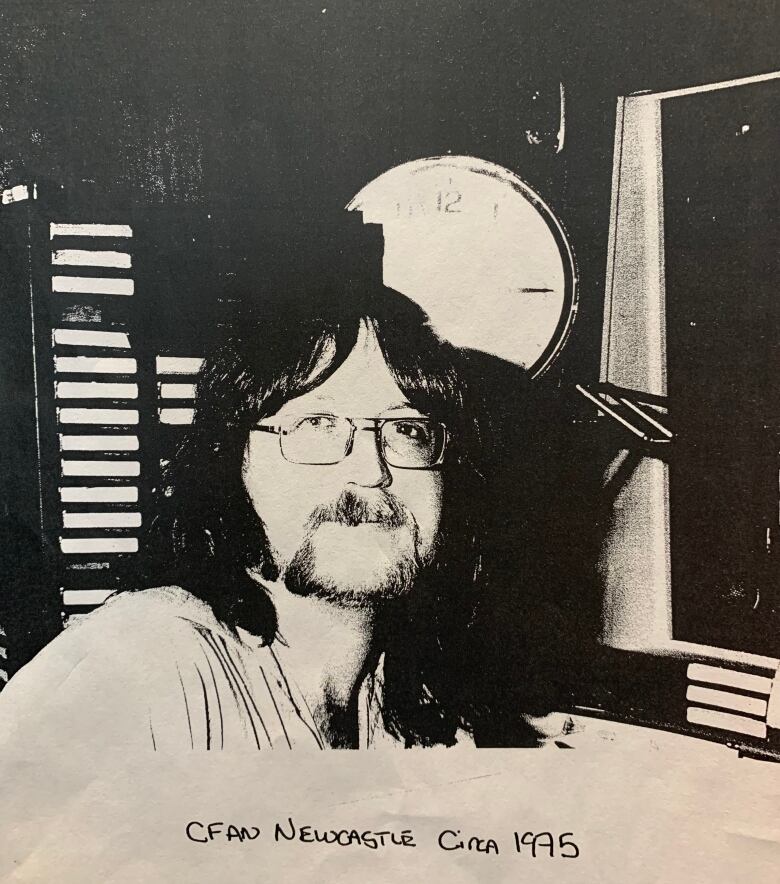 A man with long hair sits in a radio booth in a black and white photo labeled 