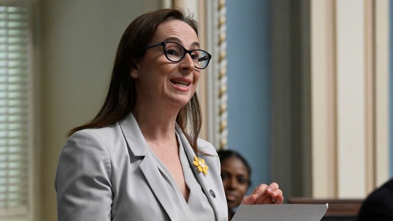 A bespectabled woman with long hair and a blazer reads from a piece of paper in the legislature.