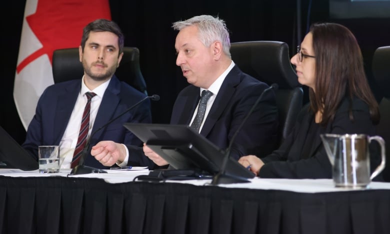 Two men and a woman wearing suits sit at a table in front of a Canadian flag.