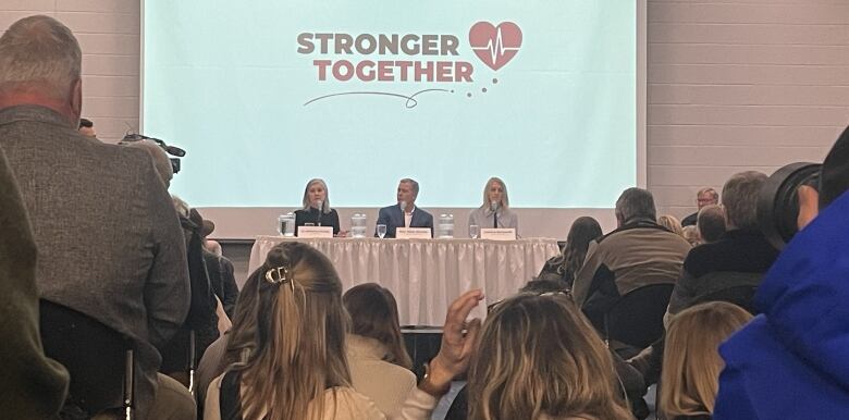 Three people sit at a table in front of a crowded conference room under a projection displaying the words stronger together