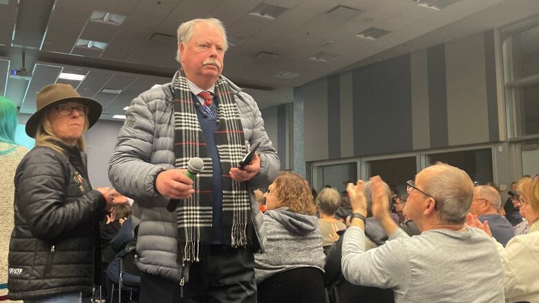 Grey-haired man with mustache, in grey puffer jacket and plaid scarf, is applauded by members of the audience as he carries a microphone. 