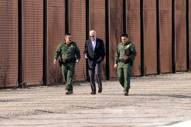 Biden in suit without tie, walking along rust-coloured border wall, flanked by agents in green uniforms