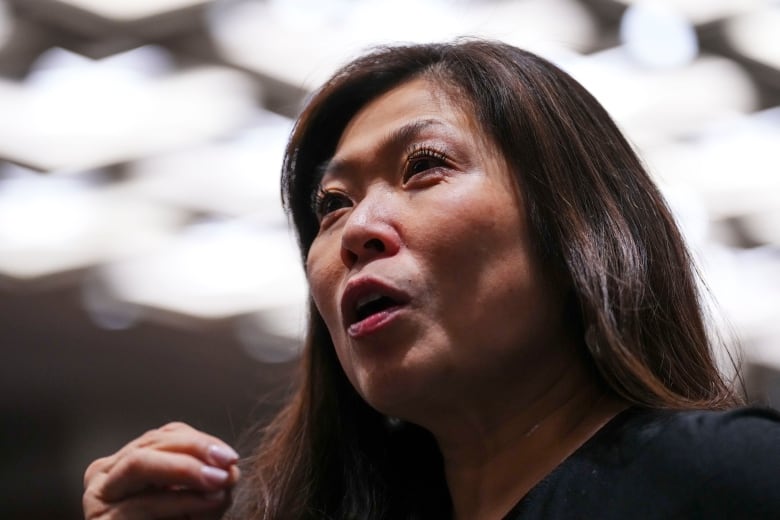 A woman with black hair wearing a black blazer gestures as she speaks into a microphone.