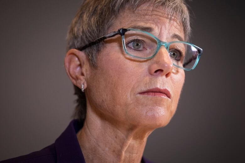 A woman with grey hair a teal-coloured glasses looks on during a news conference.