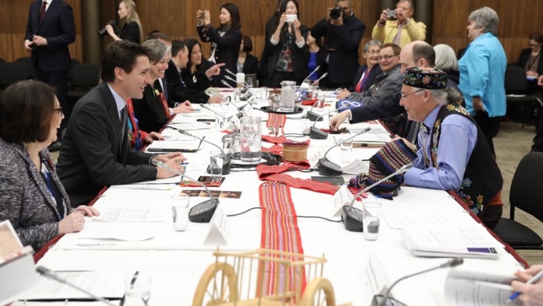 Politicians laugh and talk around a table covered with papers.