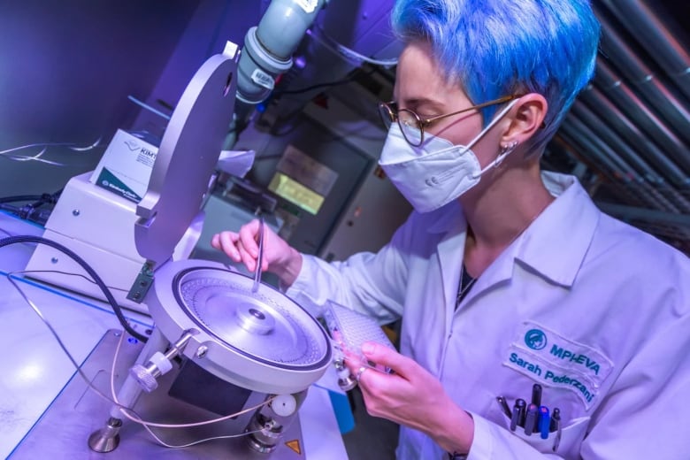 A woman with short blue hair and glasses is in a lab. She looks over a metal machine, loading small fragments inside the machine with tweezers.