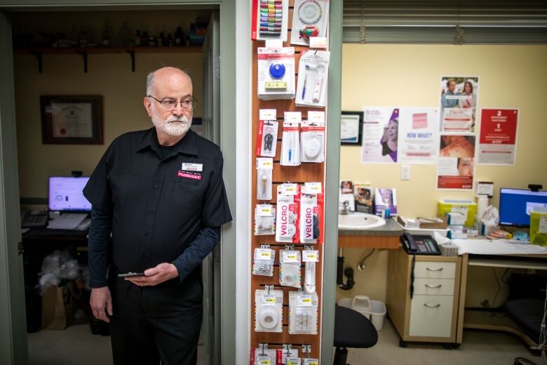 A white male with a white beard stands in the doorway of a pharmacy office and holds a cellphone.