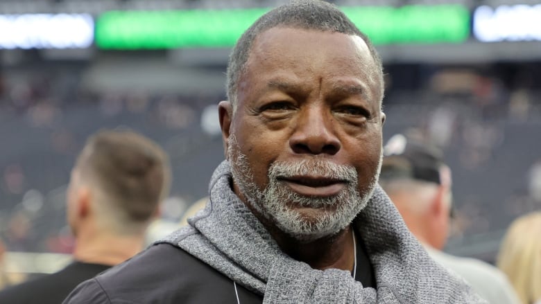 A man with a grey sweater folded around his neck smiles slightly at the camera. 