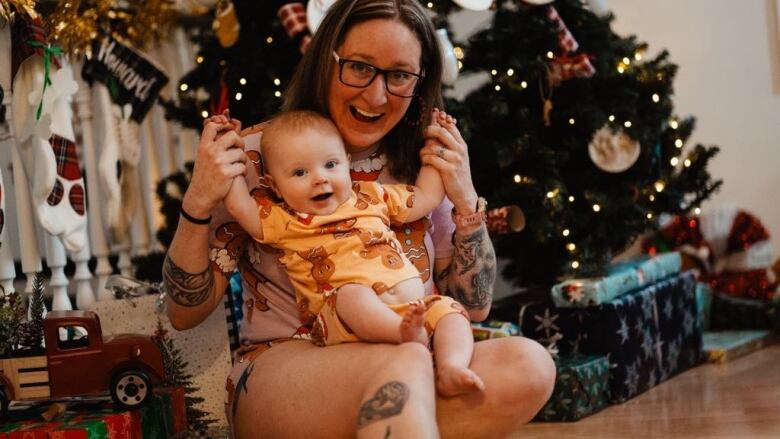 The photo shows a woman with a young baby on her laps in front of a christmas tree, laughing together.