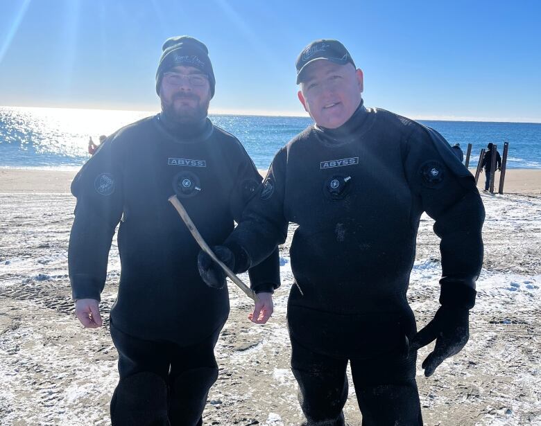 Two men in wetsuits stands next to each on a sunny day. One man is holding copper pipe about the size of an axe handle.