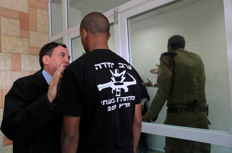 Two men stand in front of a glass door talking.