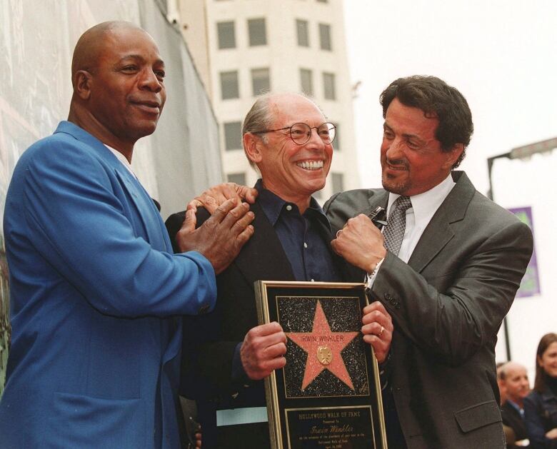 Three men smile and pose for a picture, with the one in the centre holding a framed star.