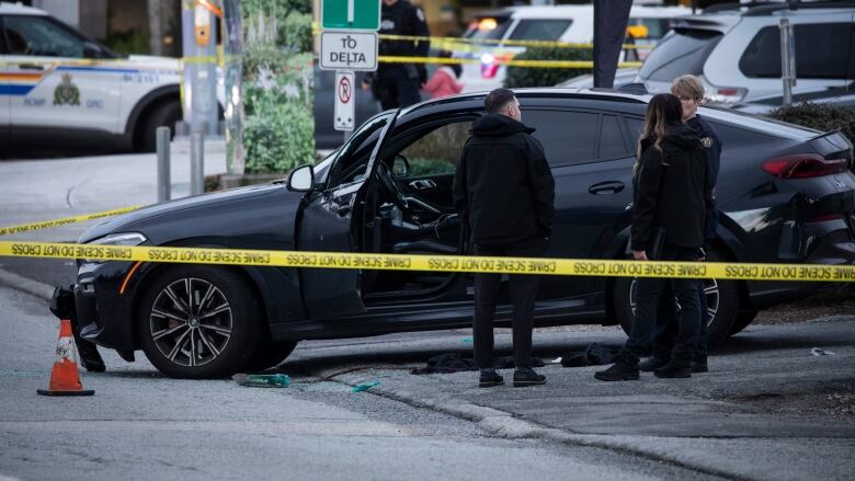 Police officers mill around behind yellow crime tape. A black car is pictured with its windows smashed.