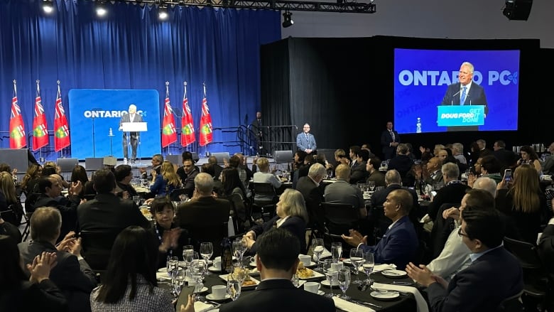 A crowd seated at dinner tables in the foreground looks toward Doug Ford as he stands on a stage in front of a sign that says 'Ontario PC.'   