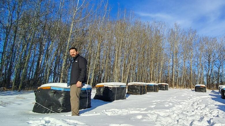 A white man with dark hair and a beard is fully clothed. He is outside on a clear sunny day, standing in the snow. Behind him are a line of boxes with black tarp tied around them.