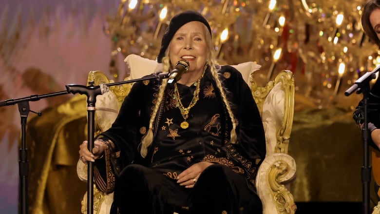 A woman wearing a black outfit with gold details sits in a chair and sings into a microphone.