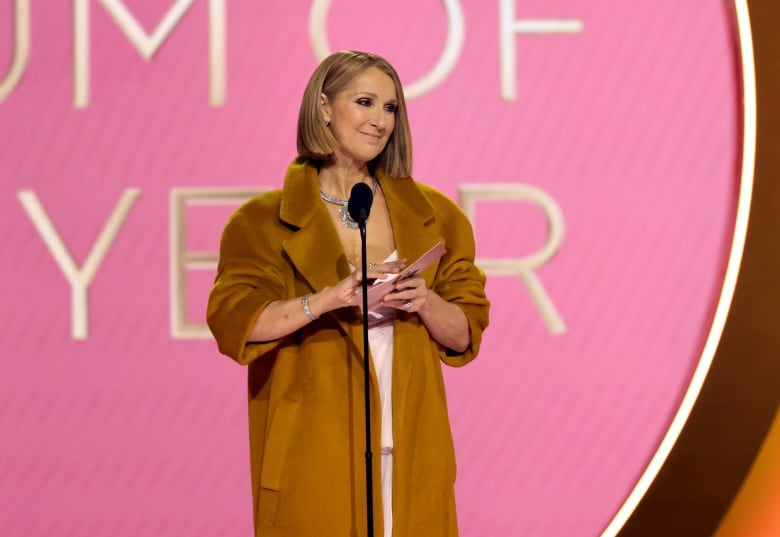 A woman wears a camel-coloured coat and a pink gown while holding an envelope.