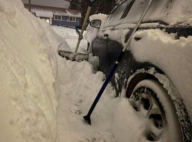 A shovel and car scraper lean on a car with a 1.5 metre drift of snow next to it.