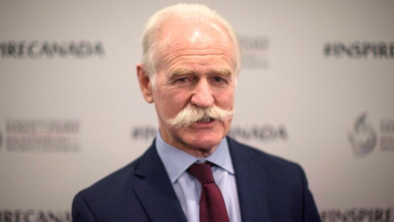 an older man with grey hair and a big moustache in a blue suit and red tie smiles for the camera