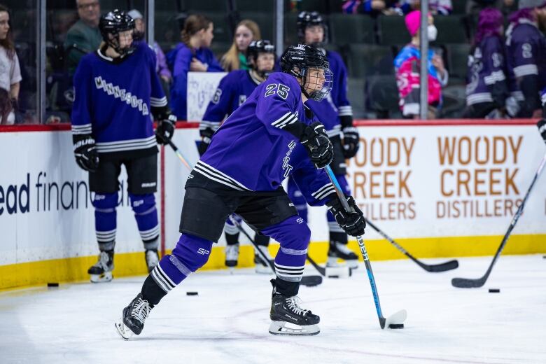 A hockey player wearing a purple jersey skates with the puck while teammates stand around in the background.