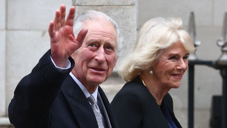 An elderly man with white hair waves as he leaves a white building with his wife, who is blonde.