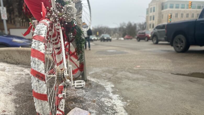 A white-pained bike wrapped with red ribbon and with flowers on a sidewalk near a busy intersection