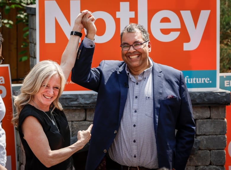 A woman raises a man's arm in celebration and points at him from below with her other arm, with an orange NDP sign behind them.