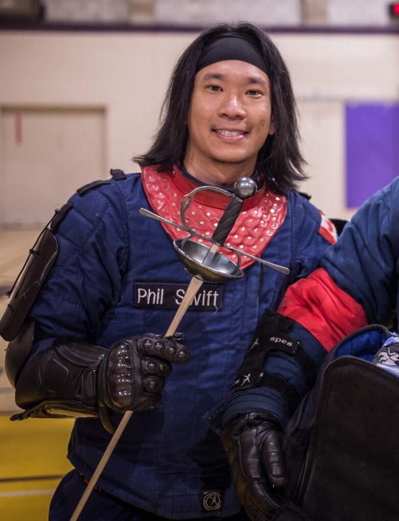 A smiling man dressed in blue protective clothing holds a sword.