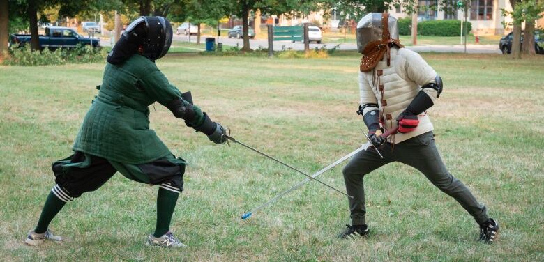 Two people, dressed in medieval style fencing attire and masks, spar outdoors on grass.