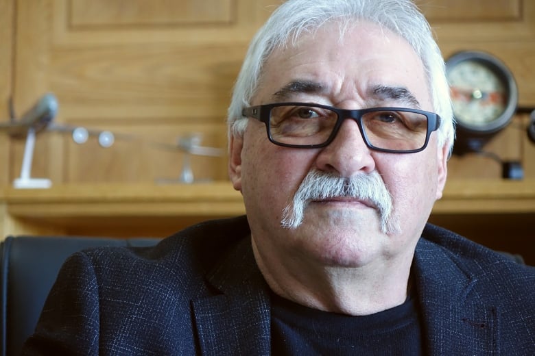 A man with gray hair and a handlebar moustache, wearing glasses, looks into the camera.