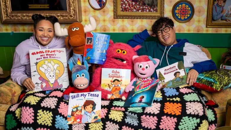 Two people and four puppets sit on a sofa under a blanket and smile at the camera. They are each holding a picture book.