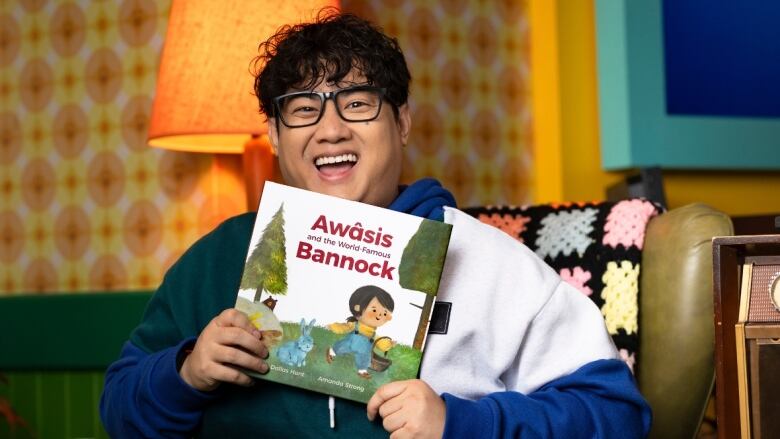 A man with dark hair, wearing glasses, holds up a picture book to the camera, smiling.