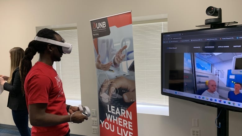 Man wearing virtual reality headset, holding controllers in his hand facing television.