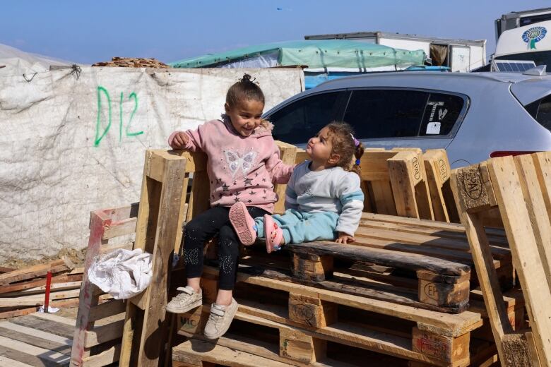 Two young girls sit on wooden pallets in front of a car. 