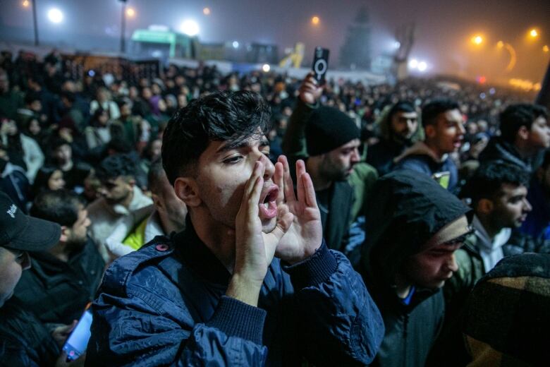 A large crows of people stand outside on a foggy night. In the foreground, a man cups his hands around his mouth and shouts. 