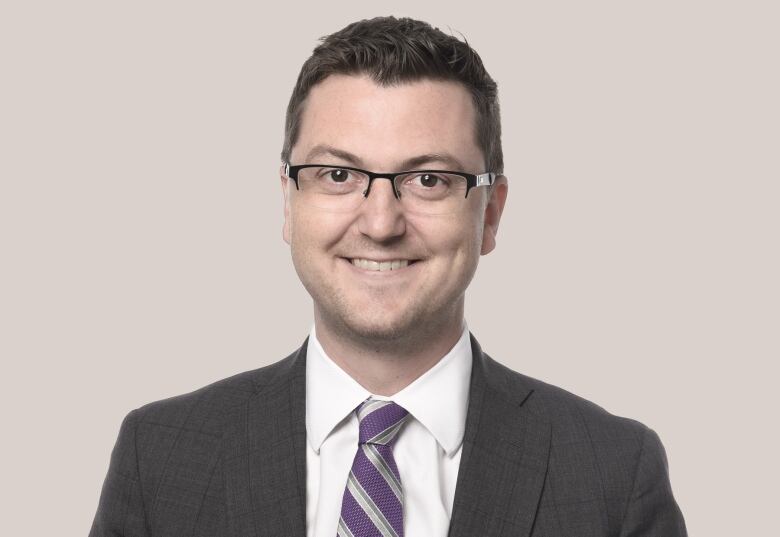 A man stands in front of a grey beige wall wearing black rimmed glasses, a white collared shirt, grey suit jacket and purple and grey horizontally striped tie.