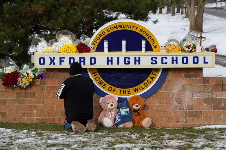 A person kneels on snow-covered grass beside a collection of stuffed animals in front of a school sign that has bouquets of flowers laid on top of it.