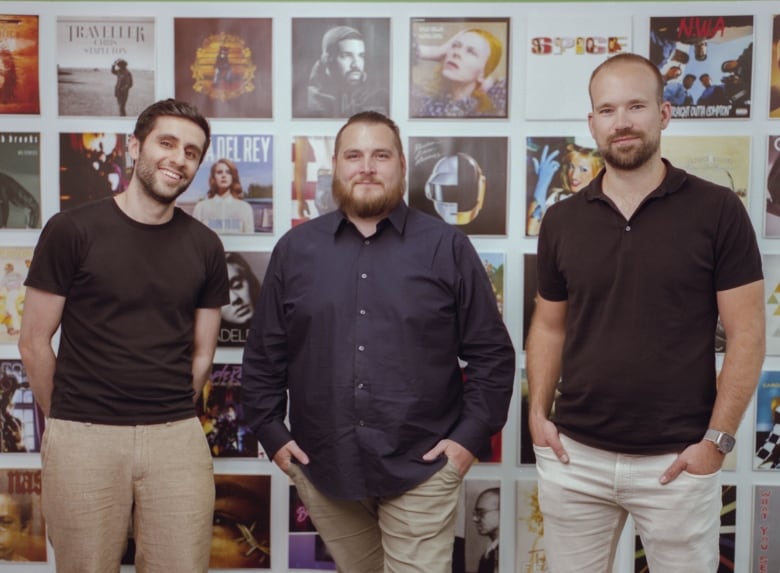 A picture of 3 white men posing in front of a wall of album covers.