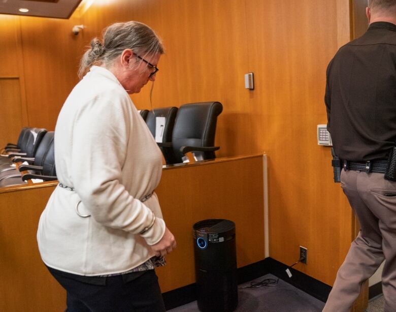 A woman wearing a white sweater and glasses walks toward out of a courtroom in handcuffs attached to a chain around her waist.