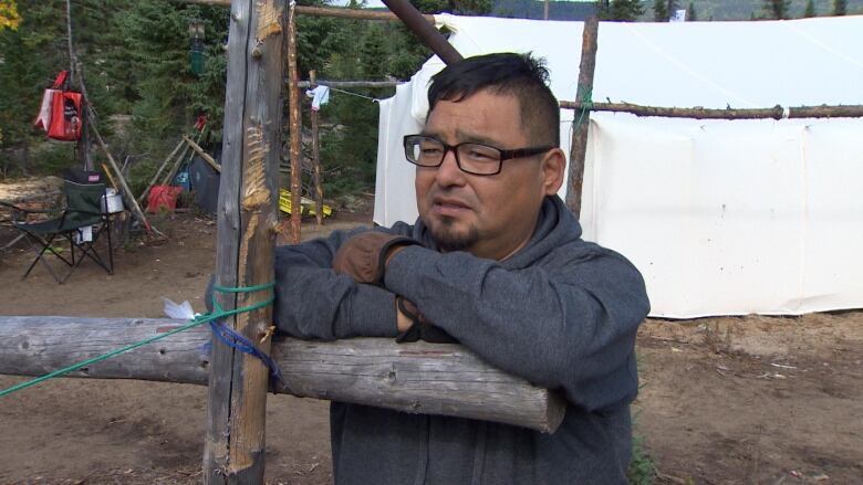 A man wearing a grey sweater leaning against a fence post. 