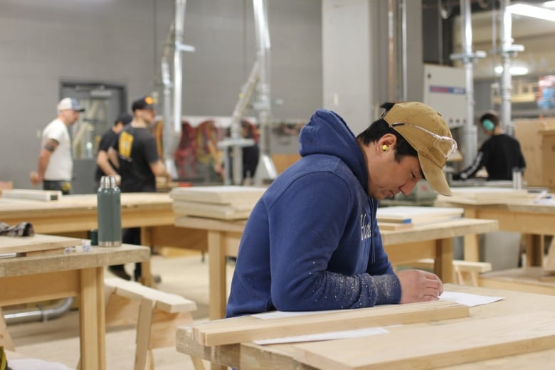 A student in a blue hoodie and baseball cap is pictured drawing a project inside a carpentry lab.