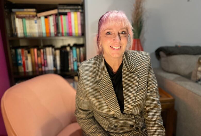 A woman with pink hair in a blazer smiles at the camera. There's a book shelf behind her.