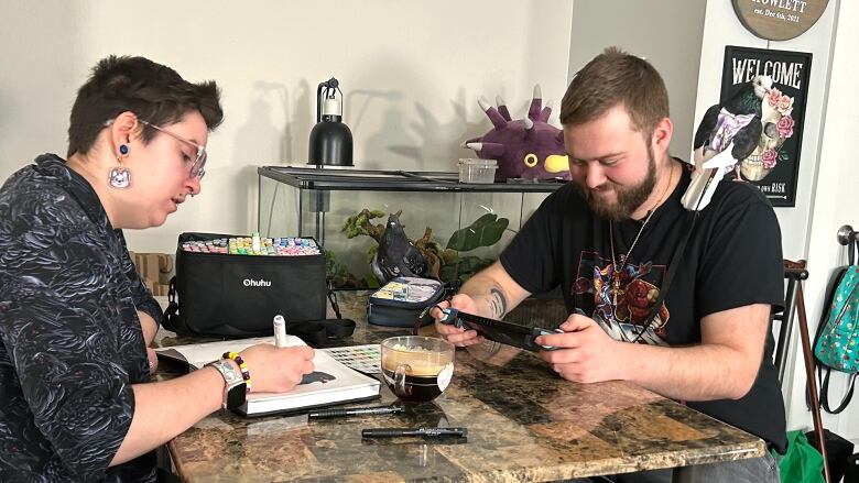 a man and a woman sit at table, drawing and playing a video game with a pigeon sitting on the man's shoulder 