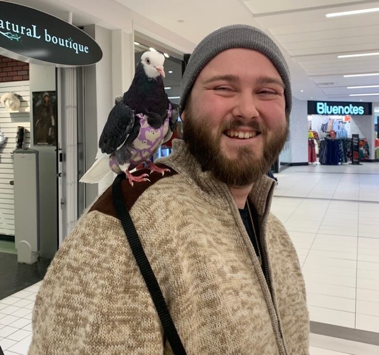 A man in a beanie stands indoors in a mall with a pigeon on a leash and in a diaper on his shoulder 